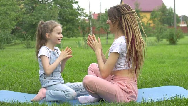 Young mother with dreadlocks hairstyle and little daughter are playing with each other while doing yoga exercises on grass in the park at the day time. Friendly family