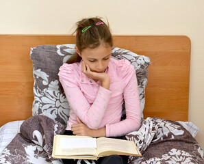 A girl reads a book, sitting in a bed and holding her head with her hand