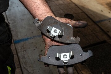 A car mechanic holds in a dirty, open hand a new and old brake pad, visible rear part of the pads.