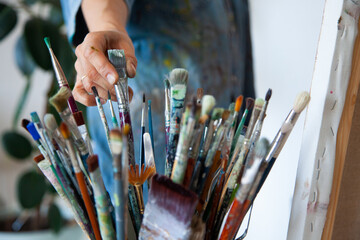 Woman artist hand choosing and picking oil paintbrushes near canvas in workshop - obrazy, fototapety, plakaty