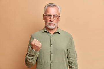 Portrait of irritated bearded mature man clenches fist and demonstrates anger warns you dressed in...
