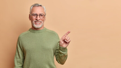 Smiling positive senior man with grey hair and beard points right and shows perfect copy space has...