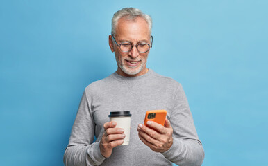 Cheerful modern senior man uses cellphone for communication types text message on phone screen...