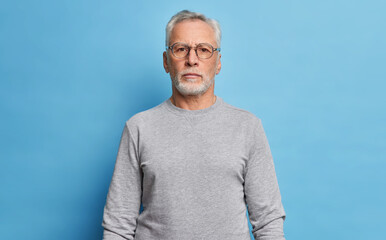 Shot of serious bearded mature European man dressed in casual grey jumper looks directly at camera being confident in himself poses against blue studio background. Handsome grandfather on pension - obrazy, fototapety, plakaty