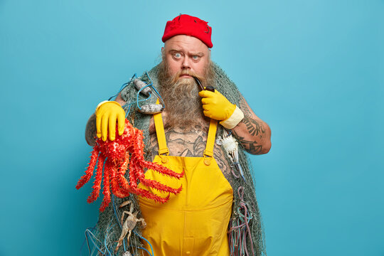 Indoor shot of serious bearded sailor raises eyebrows and looks strict at camera has tattooed body dressed in yellow overalls red hat with fish hooks carries fishing net smokes pipe holds octopus