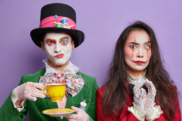Photo of serious scary woman with painted bloody scars on face steepls fingers with intention to do something. Funny man hatter has colorful makeup dressed in costume drinks tea. Halloween theme