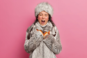 Indigenous circumpolar woman yawns and has sleepy expression wears winter clothes lives at greenland poses against rosy background. Tired Nomadic nenet female dressed in grey fur coat and hat