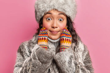 Cropped shot of astonished winter woman in warm outerwear stares at camera surprisingly keeps hands on face poses against pink background. Shocked eskimo girl wears traditional nothers clothes - obrazy, fototapety, plakaty