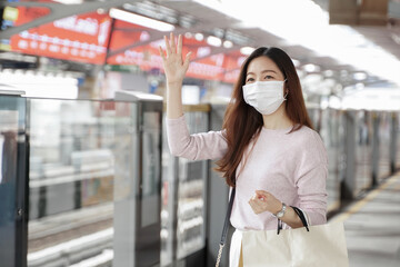 Asian business woman in casual clothes wearing face mask raised her hand to greet friends while waiting for the train to go to work on the platform station. New normal lifestyle in city concept.