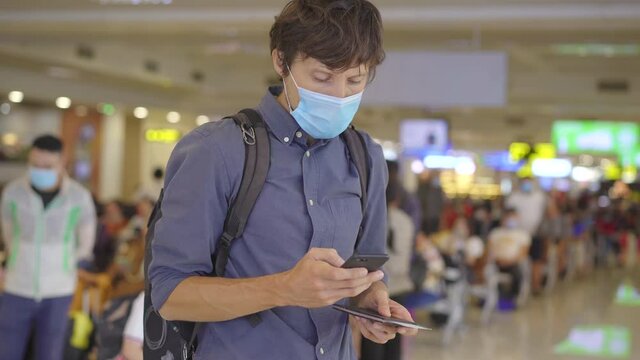 A young man wearing a medical face mask in an airport stands in a hall with a smartphone in his hands. The concept of the New normal of people's lifestyle. Air travel in the age of Covid-19