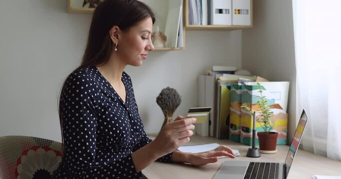 Smiling focused young beautiful european woman holding bank credit card, entering payment information cvv code, approving money transaction in computer shopping application or e-banking system.
