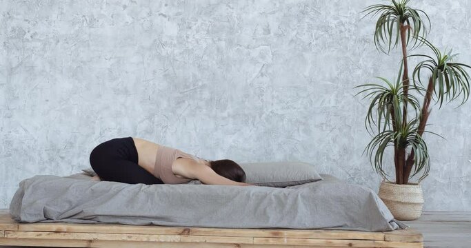 Young fitness woman brunette in sportswear practicing yoga on bed in bedroom in morning. Slim girl with sexy bod doing exercise lazy workout raises leg up strains muscles falls from fatigue on blanket