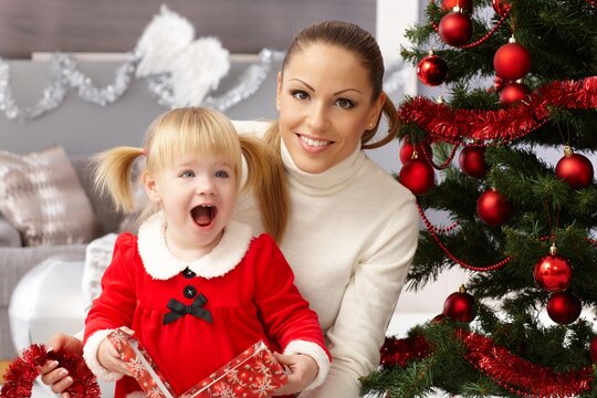 Happy little girl holding christmas surprise sitting by christmas tree with mother. 