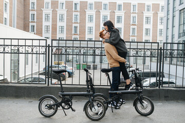 Ginger young man with glasses holding his girl in the air by her thighs in front of the fence. Ebikes in foreground. Side view. Far wide shot. Over high storey building in a background.