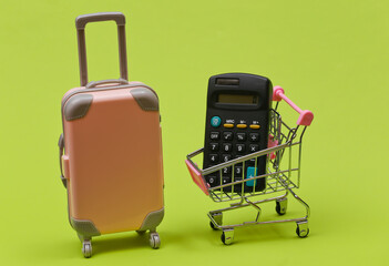 Travel and shopping concept. Mini plastic travel suitcase and supermarket trolley with calculator on green background.