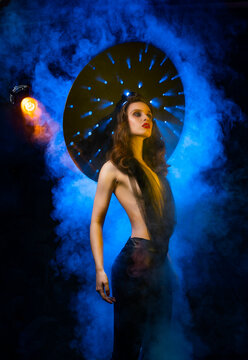 A beautiful slim sexy girl with evening make-up and red lips, wearing a large hat with holes through which rays of light shine, posing in colorful smoke. Artistic, conceptual, advertising design