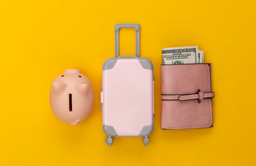 Travel flat lay. Mini plastic travel suitcase, purse and piggy bank on yellow background. Minimal style. Top view