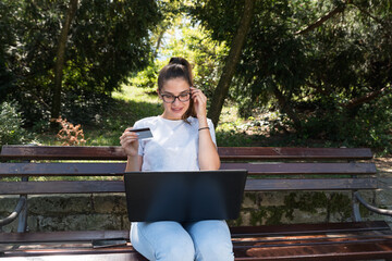Obraz premium Happy young woman or girl sitting in the park with laptop and bank credit card and smiling after successful online shopping 