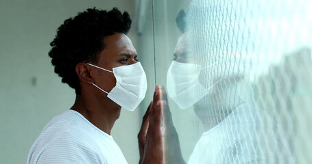 African man holding into window with hands during quarantine lockdown wearing virus mask