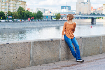 An adult red-haired woman in casual clothes stands on the embankment, leaning against a stone fence and turning in surprise towards the water. River embankment on a warm day.