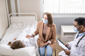 Young woman in protective mask sitting by bed of sick friend in covid hospital
