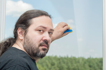 A disgruntled adult man with a displeased face and beard washes a window with a sponge at home. Man on a light background.