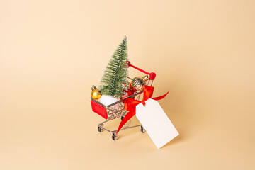 Mini shopping cart with artificial Christmas tree, red and golden Christmas balls, tag with red ribbon on a beige background. New year concept. Idea for christmas sale, shopping