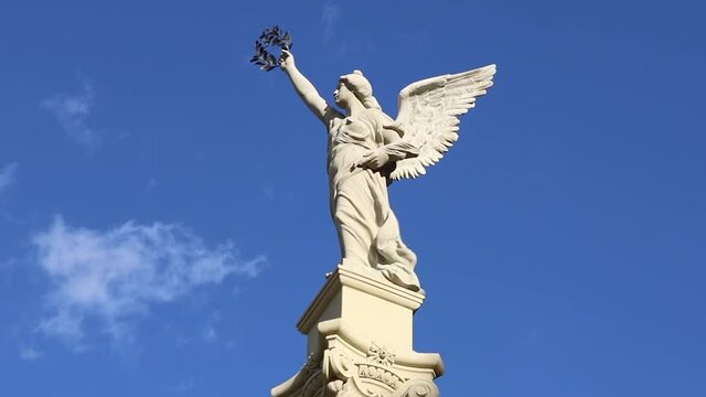 Statue of a female angel with wings and a laurel wreath in the hand, with a blue sky in the background and clouds - timelapse with the peace symbol