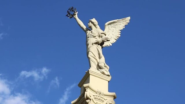 Statue of a female angel with wings and a laurel wreath in the hand, with a blue sky in the background, clouds and birds flying - peace symbol of the heaven