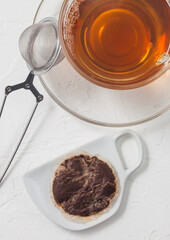 Used teabag on ceramic plate with glass of tea and strainer infuser on white.