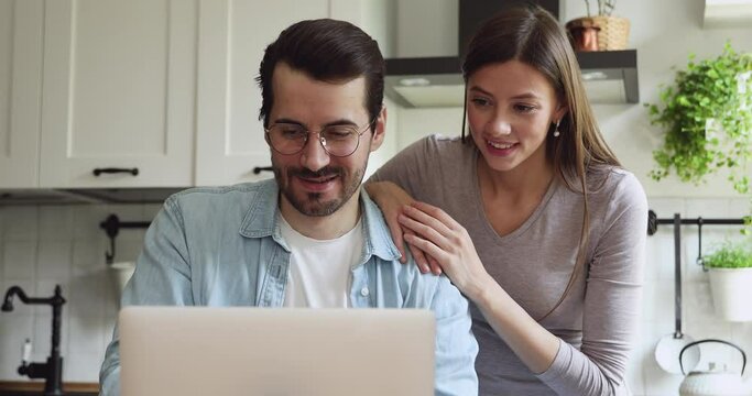 Happy young family couple watching funny video on laptop, web surfing information online, planning vacation booking flight hotel tickets, using computer software application together at home.