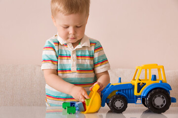 A blond boy in striped football plays at home in the children's room with colorful cubes and a large bright tractor on the table.