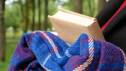 A book with a blue checkered woolen blanket or plaid in the hands of a woman wearing a sweater and black coat in the park. Warm and sunny weather. Soft, cozy photograph. Close-up
