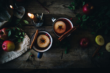 Flat lay with two metal cups with aromatic apple and cranberries tea with cinnamon stick, burning candles, apples and fir branches on old wooden table.