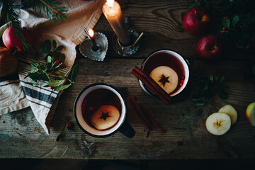 Flat lay with two metal cups with aromatic apple and cranberries tea with cinnamon stick, burning candles, apples and fir branches on old wooden table.