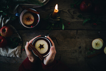 Woman hands holding one of  two metal cups with aromatic apple and cranberries tea with cinnamon stick, burning candles, apples and fir branches on old wooden table.