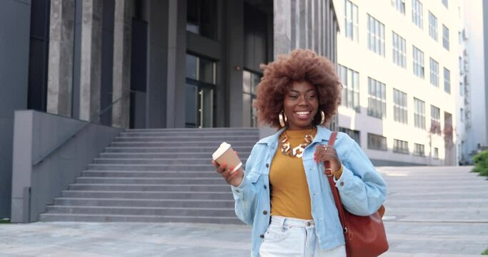 Young stylish African American beautiful curly woman with bag walking at city street, holding cup of coffee-to-go and smiling joyfully. Pretty happy female strolling outdoor with coffee and laughing.