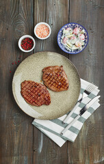 Two grilled ribeye steaks, spices and tomato-cucumber salad with sour cream on a wooden table.