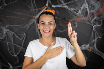 Young beautiful woman over black background with cobwebs and spiders smiling swearing with hand on...