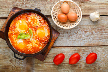 israeli fried eggs shakshuka with cheese in a frying pan near eggs and tomatoes.