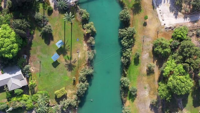 Aerial pass over Kibbutz Nir David with Asi river channel turquoise water dividing east and west side riverside houses and palm trees, Israel.