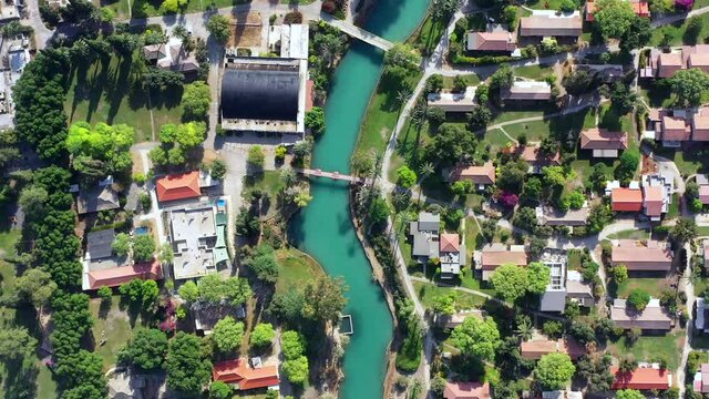 Aerial pass over Kibbutz Nir David with Asi river channel turquoise water dividing east and west side riverside houses and palm trees, Israel.