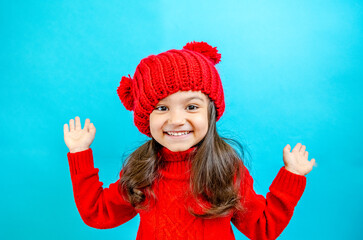 Portrait of a little curly-haired girl in a knitted red hat in winter. little girl with dark hair in a red knitted hat and sweater smiling on a blue background isolate, place for text. Winter clothes