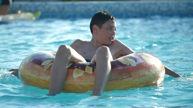 Smart brunet boy smiling and sitting on water tube in a swimming pool in slow motion