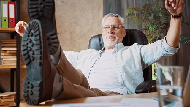 Gray-haired man putting feet on desk next to notebook and smiling leaning back in armchair, working at home