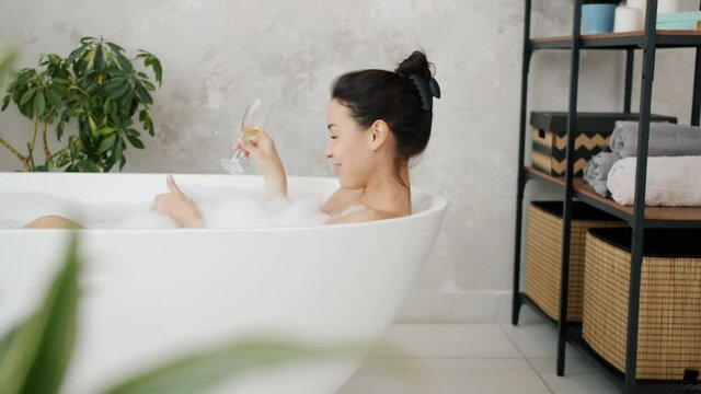 Cheerful careless lady is drinking from glass and having fun blowing soap foam in bathtub at home. Happy people and leisure activities concept.