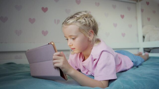 Side view of blonde little girl lying on bed at home and using digital tablet for studying or browsing Internet