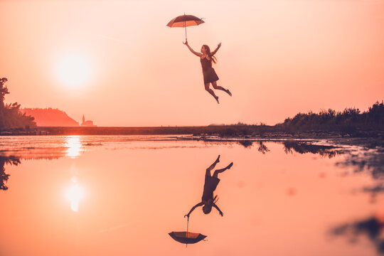 silhouette of a woman jumping with an umbrella over a sea in an orange sunset - freedom, creativity, independency and imagination concept.