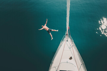 Fit caucasian white man jumping from a sailboat full body into the sea on a sunny summer day, wide...