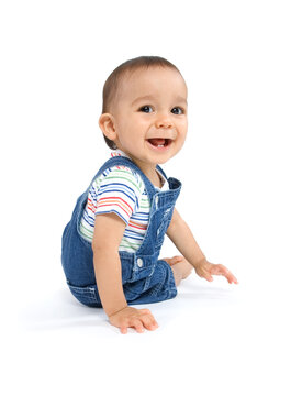 A baby boy sitting on the floor smiling at camera, dressed in overalls and isolated on white background